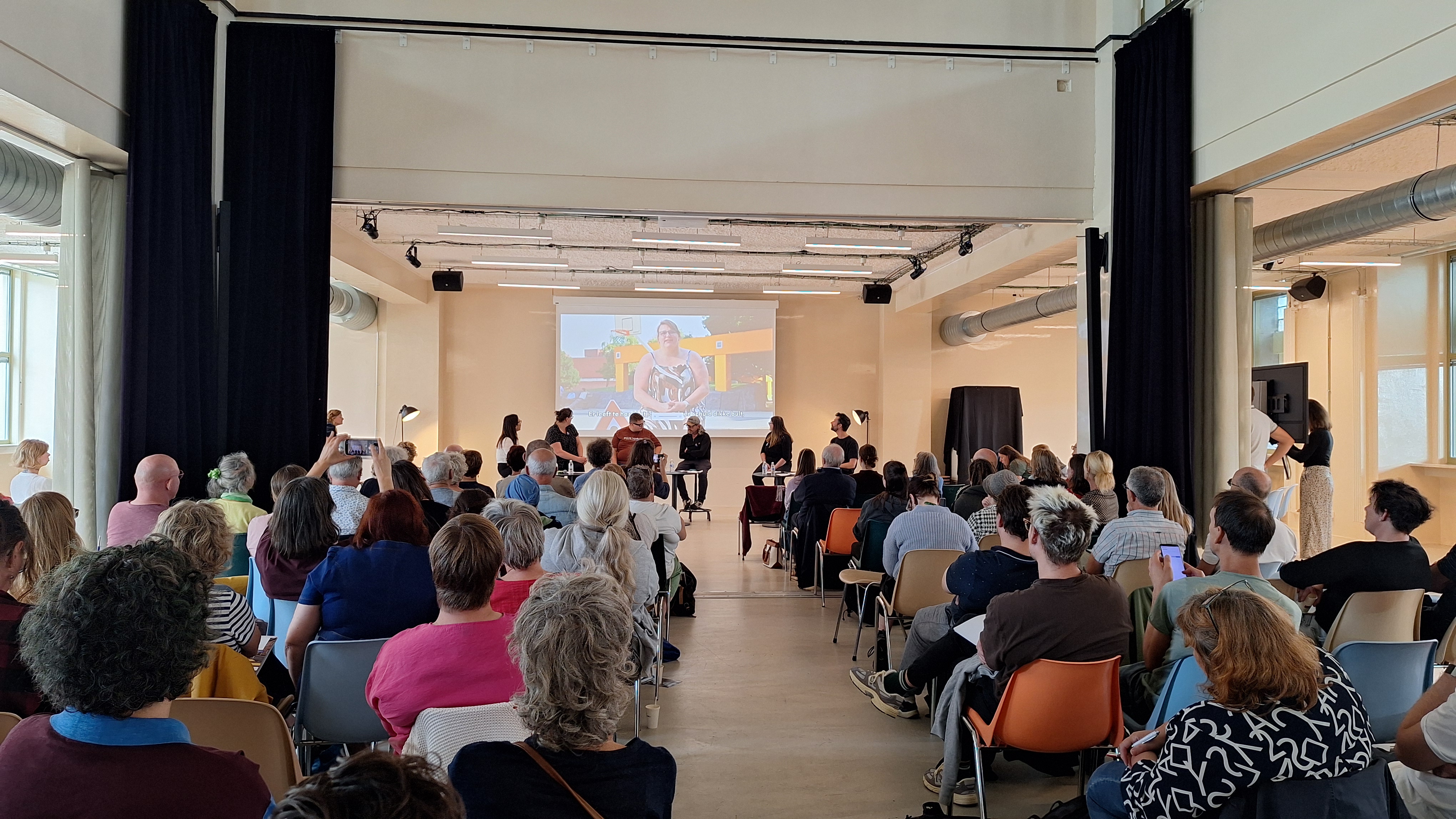 Een groep mensen luistert naar het panelgesprek vooraan in het gebouw. Vooraan zitten 6 mensen. 